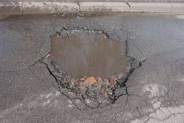Kahverengi su birikintisi olan bir çukur ve yoldaki beton kaldırım kenarındaki gri asfaltta kirli su.