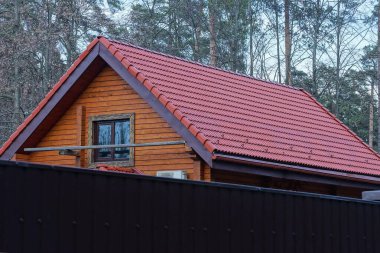 Kahverengi ahşap bir evin tavan arası. Gökyüzüne ve ağaçlara karşı bir çitin arkasındaki kırmızı kiremitli çatının altında bir pencere var.