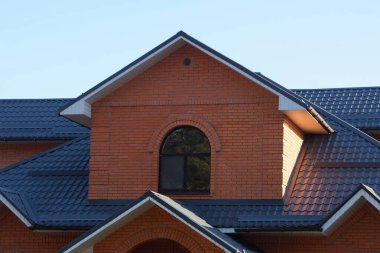 Kahverengi tuğlalı özel bir evin ön cephesi. Mavi gökyüzüne karşı döşeli bir çatının altında penceresi var.