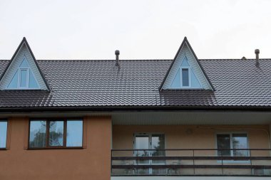 Gökyüzüne karşı kahverengi kiremitli çatının altında pencereleri olan büyük özel bir evin parçası.