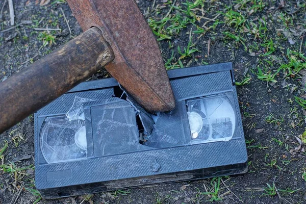 錆びた鉄のハンマーが外の地面にある古い黒いプラスチック製のビデオカセットを — ストック写真