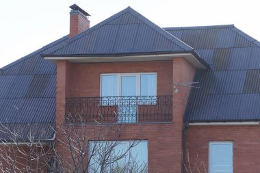 Beyaz pencereli, siyah, açık demir bir balkon ve özel bir evin kahverengi tuğlalı tavan arasında gökyüzüne karşı bir çatı altında bir kapı.