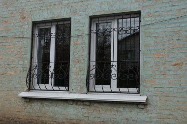 Yeşil tuğla bir duvarda işlenmiş demir desenli siyah demir parmaklıklı iki beyaz plastik pencere.
