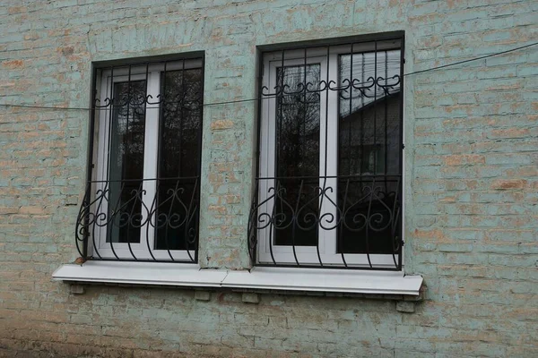 two white plastic windows with black iron bars with wrought iron pattern on a green brick wall