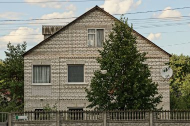 Pencereli beyaz tuğlalardan ve gri beton çitin arkasındaki yeşil ağaçtan yapılmış büyük özel bir ev.