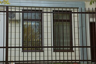 Binanın gri duvarında iki kahverengi pencere demir parmaklıklardan yapılmış siyah metal bir ızgara var.