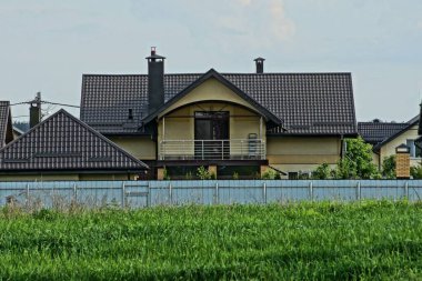 Kahverengi kiremitli çatının altında balkonu olan büyük, özel bir ev yeşil çimlerde gri metal bir çitin arkasında.