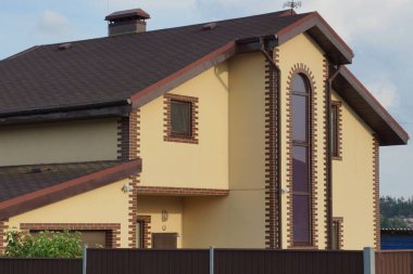 Güneşli bir günde, bir çitin arkasında mavi gökyüzüne karşı fayanslı çatı altında büyük pencereleri olan kahverengi bir evin ön cephesi.