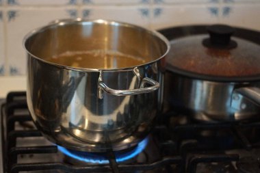 ocağın üzerinde açık gri metal bir tava duruyor ve mutfaktaki mavi bir gaz ateşi tarafından ısıtılıyor.