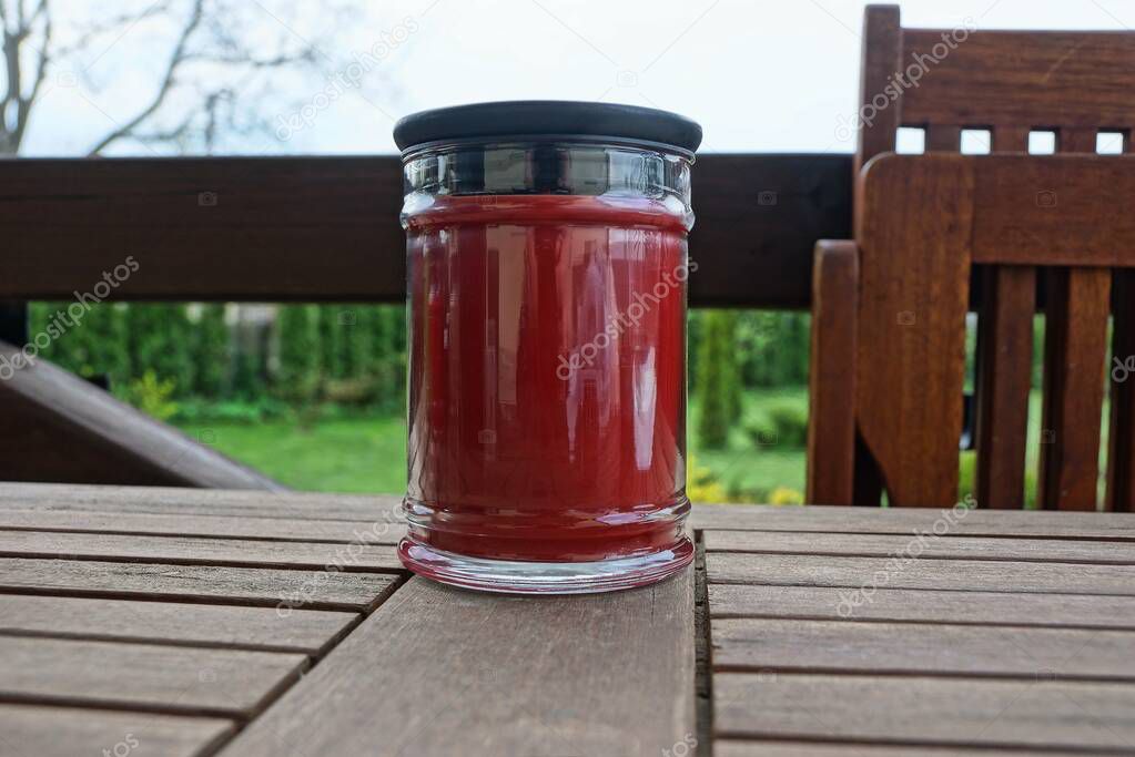 one red glass jar with a decorative candle stands on a brown board on the veranda against a background of green vegetation