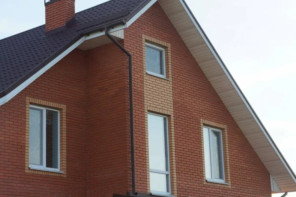 灰色の空に向かって茶色の瓦屋根の下に白い窓のある赤いレンガ造りの家の屋根裏部屋 — ストック写真