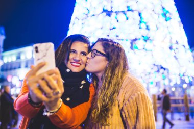 Noel ışıklarının yanında iki kız fotoğraf çekiyor. Madrid 'de birlikte eğleniyorlar. Bir tanesi arkadaşını öpüyor..