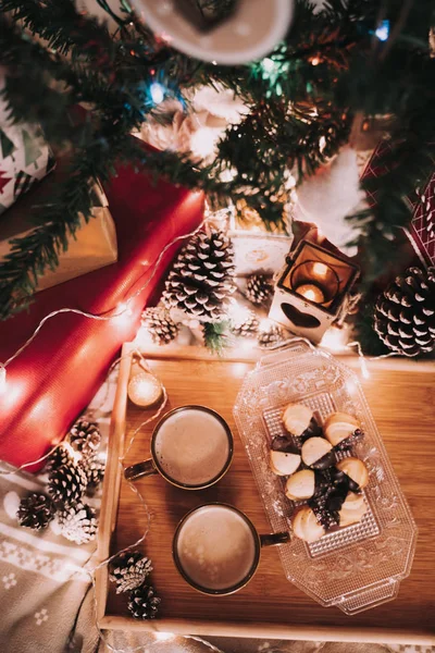 Decoración navideña con dos tazas de café y algunos pasteles en una bandeja de madera. Alrededor hay luces de Navidad, conos de abeto y algunos regalos. . — Foto de Stock