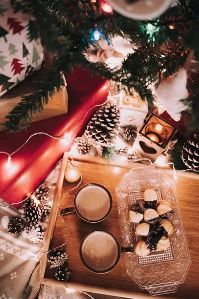 Decoración navideña con dos tazas de café y algunos pasteles en una bandeja de madera. Alrededor hay luces de Navidad, conos de abeto y algunos regalos. . — Foto de Stock