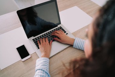 Bir iş kadını dizüstü bilgisayarına yazı yazıyor. Ahşap bir masanın üstünde. O, iş kıyafetleri giyiyor. Telefonu laptopun yanında..