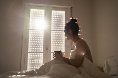A beautiful dark-haired woman is in her bed while take a cup of coffee. She is smiling. The sunlight go into the room through the window. She is cover with a eiderdown. Selective focus. clipart
