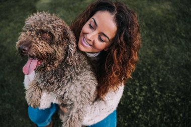 Güzel bir kadın köpeğiyle birlikte çayırda. Sahibi ona sevgiyle bakarken evcil hayvanına sarılıyor. Onlar parkta bir günün tadını çıkarıyorlar. Evcil hayvan kahverengi kürklü bir İspanyol su köpeği..