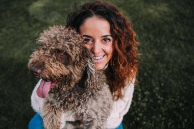 Güzel bir kadın köpeğiyle birlikte çayırda. O kameraya bakarken ev sahibi evcil hayvanına sarılıyor. Onlar parkta bir günün tadını çıkarıyorlar. Evcil hayvan kahverengi kürklü bir İspanyol su köpeği..