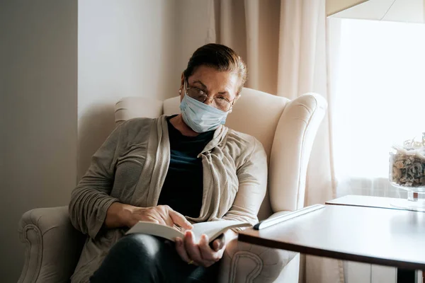 An older woman is sitting on the sofa while reading a book. She is sick and she is wearing a protective mask. She is protecting her family and herself. Pandemic concept.