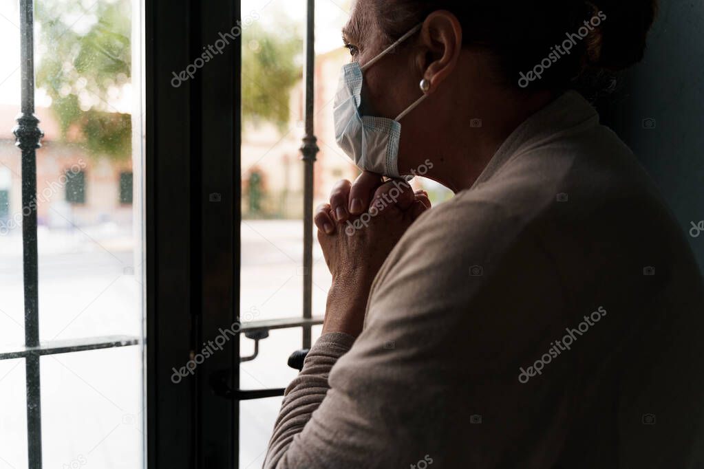 An older woman is looking through the window. She is wearing a protective mask on her face. She has her hands joined and her head is resting on them. She is protecting herself. Pandemic concept.