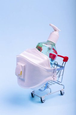 Solunum cihazı ve bir şişe alkollü el jeli dezenfektanı olan market arabası. Mavi arka planda Coronavirus dikey alışveriş kavramı.