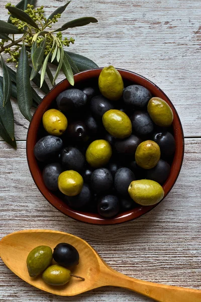 Clay bowl with black and green olives, near the olive branch and a wooden spoon