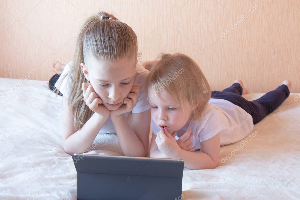 Two girls in white t-shirts on the couch with a laptop. Home schooling. Side view