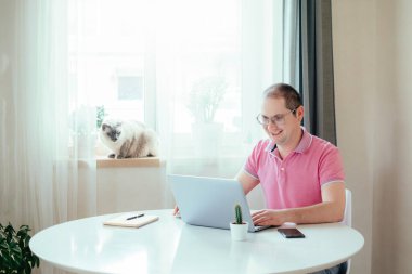 Pembe polo giyen adam masanın yanında oturuyor ve evde dizüstü bilgisayarla çalışıyor. Karantinadaki adam evden çalışıyor. Uzaktan çalışma, evden çalışma.