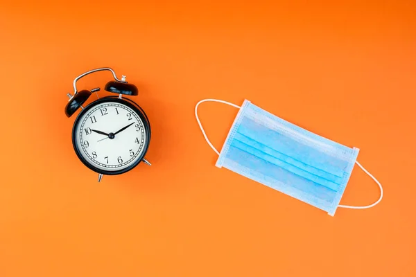 Blue face protection medical mask and black alarm clock on an orange background. Pandemic or epidemic concept.