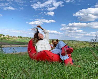 Doğanın arka planında köpeği olan bir kız ve mavi gökyüzü kırmızı bir çantanın üzerinde dinleniyor. yeşil zemin. Doğa geçmişi. Tatil kutlaması. Güzel surat. Yaz gökyüzü. İzole edilmiş kadın portresi