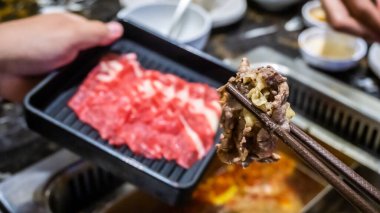 Yemek çubuğunun Japon büfe restoranındaki kaynamış çorbaya bandırılmış bir dilim eti arka plandaki bir kutuda çiğ etle birlikte tutuş görüntüsü..