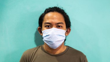 İzole edilmiş mavi arka planda 3 katmanlı cerrahi maske takan Malaylı genç bir adamın portresi. Virüsleri ve hastalıkları yaymamak için koruma adımı. Seçici odaklanma.