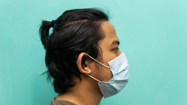 İzole edilmiş mavi arka planda 3 katmanlı cerrahi maske takan Malezyalı genç bir adamın yan görünümü. Virüsleri ve hastalıkları yaymamak için koruma adımı. Tıbbi konsept.