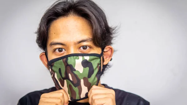 A portrait of a young Malay man wearing an army camouflage face mask on isolated white background. Protection step to avoid spreading viruses and diseases.