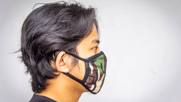 The side view portrait of a young Malay man wearing an army camouflage face mask on isolated white background. Protection step to avoid spreading viruses and diseases. Selective focused.