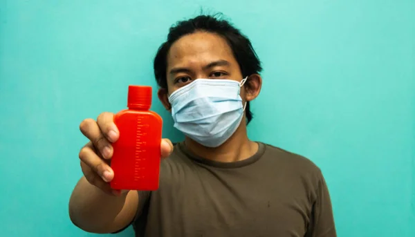 A portrait of a young Malay man wearing 3 layer surgical face mask, holding a red bottle of homemade hand sanitizer on isolated blue background. Protection step to avoid spreading virus and diseases.