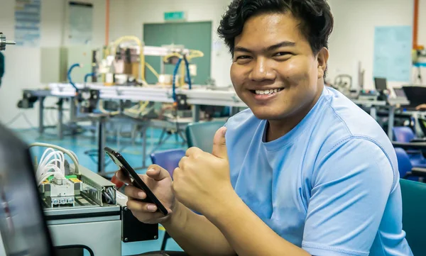 A young Malay engineering student holding a smartphone and show thumbs up with automation machine system as background at the laboratory. Image contain noise reduction. Selected focus.