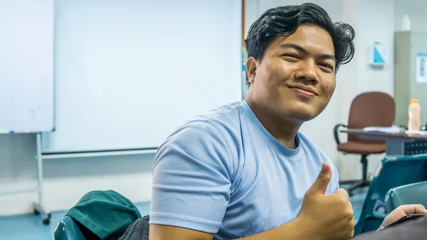Young Malay student smile brightly and showing a thumbs up or good sign in the classroom. Using a smartphone or gadget for education.