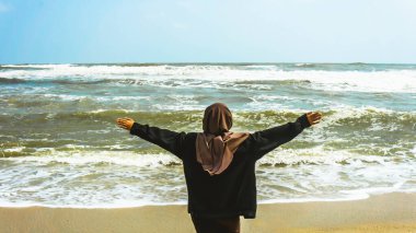 Malezyalı tesettürlü bir kızın arkadan görünüşü kollarını kaldırdı ve yaz boyunca tropik Batu Burok Sahili 'nde yürüyüş yapan okyanusa baktı. Görüntü biraz bulanık.