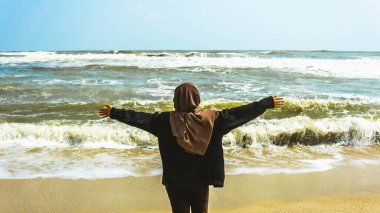 Malezyalı tesettürlü bir kızın arkadan görünüşü kollarını kaldırdı ve yaz boyunca tropik Batu Burok Sahili 'nde yürüyüş yapan okyanusa baktı. Görüntü biraz bulanık.