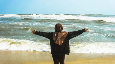 Malezyalı tesettürlü bir kızın arkadan görünüşü kollarını kaldırdı ve yaz boyunca tropik Batu Burok Sahili 'nde yürüyüş yapan okyanusa baktı. Görüntü biraz bulanık.