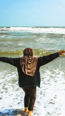 Malezyalı tesettürlü bir kızın arkadan görünüşü kollarını kaldırdı ve suyla oynadı, yaz boyunca tropik Batu Burok Sahili 'nde yürüyüş yaptı. Görüntü biraz bulanık.