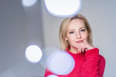 Kırmızı kazaklı kadın elinde Noel çelengi tutuyor. Ön planda çelenk ışıkları.