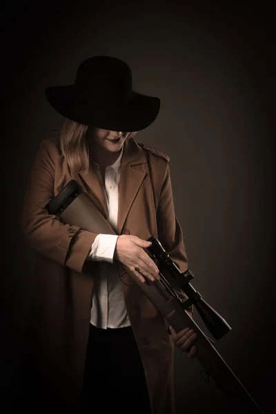 Une femme vêtue d'un manteau marron et d'un chapeau noir avec des pistolets à air dans les mains regarde à la vue. Séance photo style gangster. Fond flou foncé, foyer flou, bruit — Photo