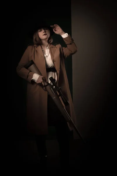 复古黑社会的女人穿着棕色斗篷手拿着气枪戴着黑色帽子用黑手党的风格拍照。黑暗模糊的背景模糊的焦点噪音. — 图库照片
