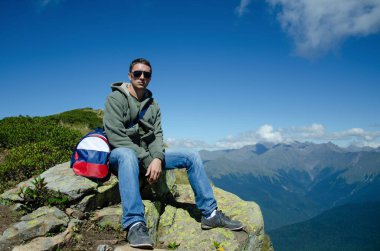 A man in sunglasses and jeans sits on top of a mountain, against the backdrop of the Caucasus Mountains with a bag on his shoulder with the image of the Russian flag.