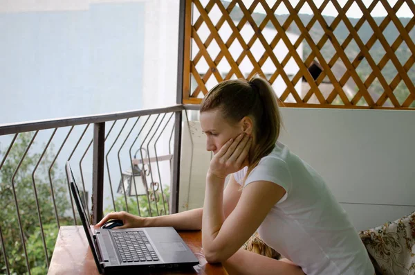 A woman in a white T-shirt on the balcony of the hotel on vacation working on a laptop. — Stock Photo, Image