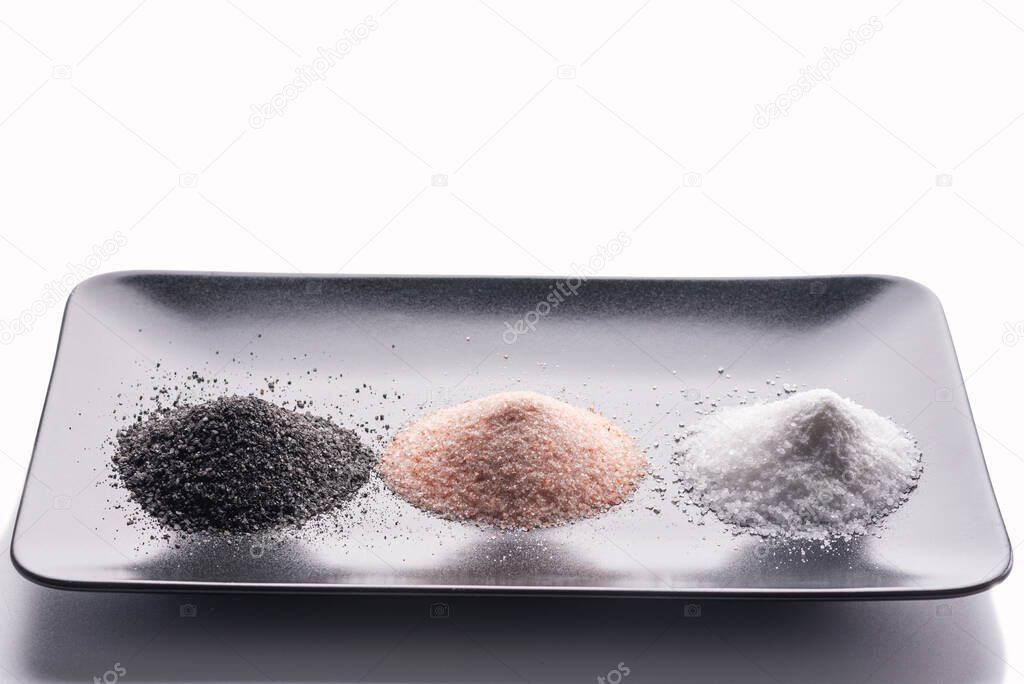 on a black plate are three types of salt - black, pink and white. On a white background.