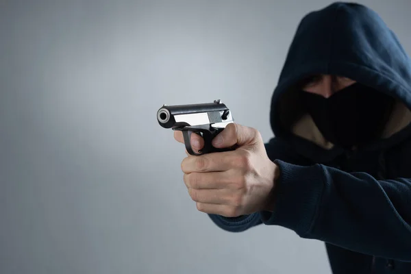 Mascarado de capuz com uma arma nas mãos sobre um fundo preto. lugar para txt em um fundo cinza — Fotografia de Stock