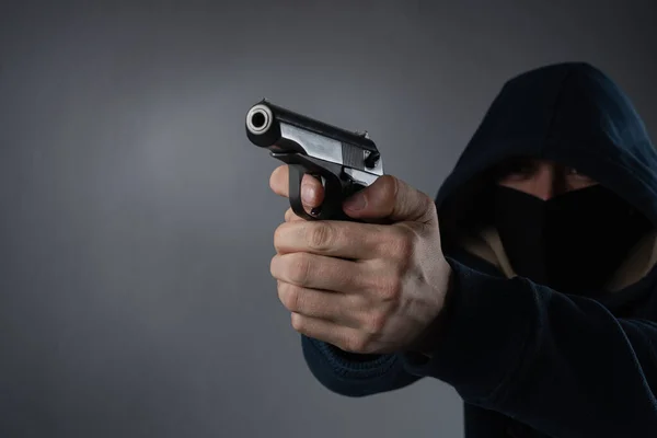 Mascarado de capuz com uma arma nas mãos sobre um fundo preto. lugar para txt em um fundo cinza — Fotografia de Stock